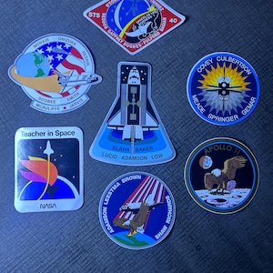 May include: A collection of six space-themed stickers featuring NASA missions. The stickers are round, square, and diamond-shaped and feature images of space shuttles, astronauts, and the American flag. The stickers are printed on white paper with colorful designs.