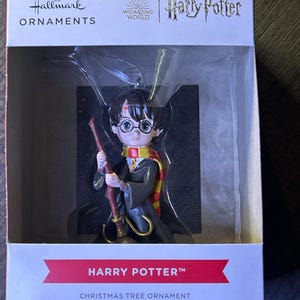 May include: A Hallmark Harry Potter Christmas tree ornament in its original packaging. The figurine depicts a young character with round glasses, a lightning bolt scar, a black robe, and a red and yellow scarf, holding a brown broomstick.