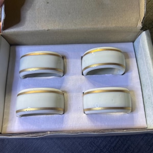 Può includere: Set di quattro anelli portatovaglioli in ceramica bianca con una fascia dorata. Gli anelli portatovaglioli sono in una scatola di cartone con interno bianco. Design classico.