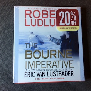 Puede incluir: Una portada de libro de audio para "The Bourne Imperative" de Eric Van Lustbader. La portada presenta una foto de dos hombres en un entorno nevado, uno apuntando con un arma al otro. El libro es una nueva novela de Jason Bourne y está narrada por Holter Graham. La portada también incluye una pegatina de venta con un 20% de descuento.