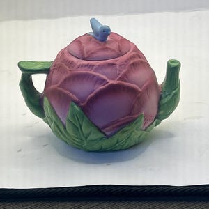 May include: A ceramic teapot shaped like a flower, with a purple body and green leaves. A small blue bird sits atop the lid. The teapot has a handle and spout, and is designed to look like a blooming flower.