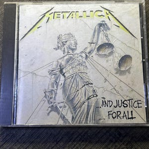 以下が含まれることがあります： 透明なプラスチックケースに入ったメタリカの音楽CD。カバーアートには、目隠しをしてロープで縛られ、天秤と剣を持った正義の女神のグレー調のイラストが描かれており、METALLICAおよび...AND JUSTICE FOR ALLというテキストが記されています。