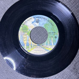 Peut inclure: Disque vinyle noir avec une étiquette colorée comportant le texte "Alice Cooper", "You and Me", "WBS 8349" et "3:25".