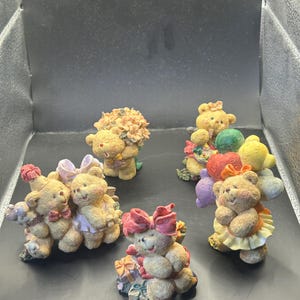 May include: A collection of small, beige teddy bear figurines. Each bear has a unique design, including bows, flowers, and gifts. Some bears are holding hearts and balloons. The figurines are arranged on a dark surface.