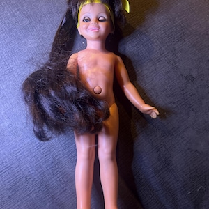 May include: A vintage doll with long, dark brown hair and a yellow headband. The doll has a smiling expression, painted facial features, and is wearing no clothing. The doll's arms are outstretched, and it is standing.
