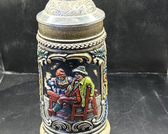 May include: A decorative beer stein with a detailed relief scene of two people. The stein has a silver-colored lid and a handle. The body of the stein is cylindrical with a detailed relief design, featuring a man and woman. The stein is approximately 8 inches tall.