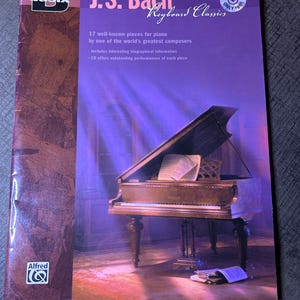 Puede incluir: Un libro morado titulado "J.S. Bach Keyboard Classics" con un piano de cola. La portada presenta texto y un logotipo. Contiene 17 piezas para piano del compositor, información biográfica y un CD.