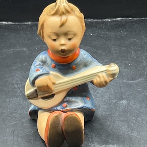 May include: A figurine of a child playing a stringed instrument. The child is wearing a blue dress with red accents and a red collar. The figurine has a light skin tone and closed eyes, suggesting it is singing.