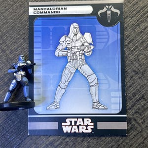 May include: A Star Wars Mandalorian Commando action figure and trading card. The figure is blue and silver, standing on a black base. The card features a detailed illustration of a Mandalorian Commando with the Star Wars logo.