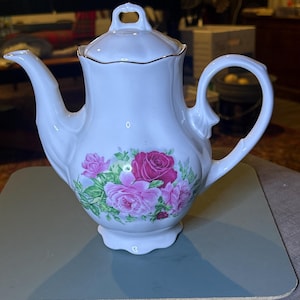 May include: White porcelain teapot with a floral design featuring pink and red roses. The teapot has a gold trim around the lid and a delicate handle.