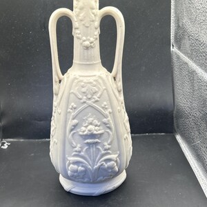 May include: A cream-colored, vintage-style vase with two handles. The vase features intricate floral and leaf patterns, with a flared top and a rounded base. The design is symmetrical, with the handles arching gracefully from the neck.