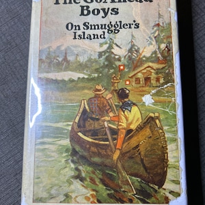 Puede incluir: Una portada de libro vintage con una ilustración de dos niños en una canoa en un lago. El título del libro es "The Go Ahead Boys On Smuggler's Island".