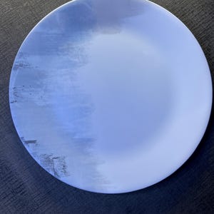 May include: A round, white plate with a blue watercolor design. The plate has a gradient effect, transitioning from a darker blue on the left to a lighter blue on the right. The plate is made of ceramic.