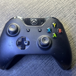 Puede incluir: Un mando de videojuegos inalámbrico negro con dos joysticks analógicos, una cruceta y cuatro botones de acción etiquetados como A, B, X e Y en verde, rojo, azul y amarillo. El dispositivo incluye un botón central con el logo de Xbox y botones de menú.