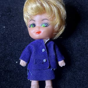May include: A small plastic doll with blonde hair, green eyes, and a purple dress. The doll is wearing white shoes.