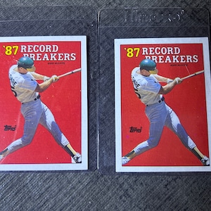 Pode incluir: Dois cartões de beisebol com um jogador de beisebol com um uniforme branco balançando um taco. Os cartões são da série Topps Record Breakers de 1987 e apresentam Mark McGwire. Os cartões estão em mangas de plástico protetoras.