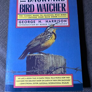 Op de afbeelding: De omslag van het boek "The Backyard Bird Watcher" toont een vogel met een gele borst die op een houten paal zit tegen een blauwe lucht. De titel van het boek staat in grote, vetgedrukte letters en de naam van de auteur staat eronder. Een citaat van John Strohm, redacteur van National/International Wildlife, staat onderaan.