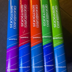 May include: A collection of five Augsburg Sermons books. The books have colorful spines in blue, red, orange, green, and lime green. Each book has the title "Augsburg Sermons" in white text. The books are labeled "Gospels Series A", "Gospels Series B", "Gospels Series C", "Epistles Series B", and "Epistles Series C".