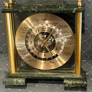 May include: A rectangular quartz world time clock featuring a dark green marble base and top, supported by four gold-toned metal pillars. The circular gold-toned face displays a central map, a 12-hour dial, a 24-hour ring, and various global city names.