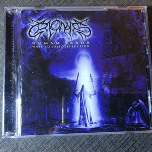 May include: A CD case with a dark blue and purple background. The band name "Crionics" is written in a gothic font. The album title "Human Error Ways to Self-Destruction" is written in white text. A figure in a white robe stands in the center of the image, holding two swords.