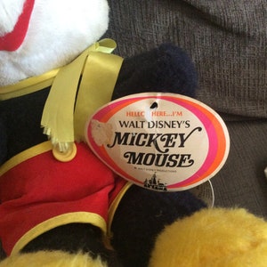 May include: Vintage Walt Disney's Mickey Mouse plush toy with a yellow ribbon around its neck. The toy is wearing red shorts with yellow trim and a black and white shirt. The toy has a tag that reads "Hello there...I'm Walt Disney's Mickey Mouse".