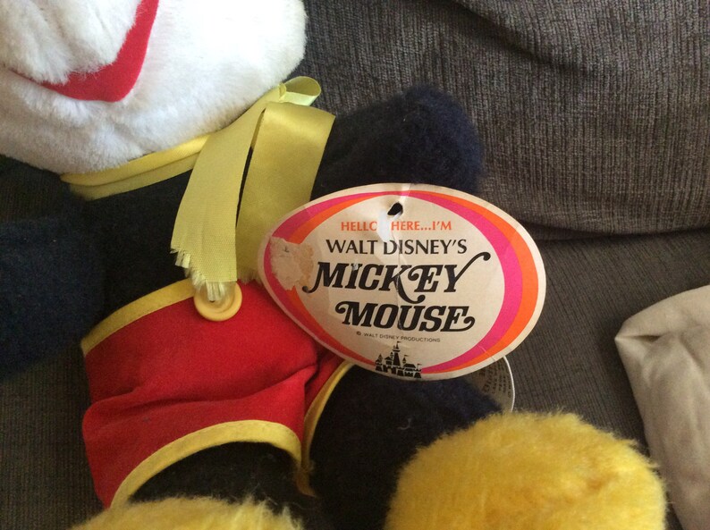 May include: Vintage Walt Disney's Mickey Mouse plush toy with a yellow ribbon around its neck. The toy is wearing red shorts with yellow trim and a black and white shirt. The toy has a tag that reads "Hello there...I'm Walt Disney's Mickey Mouse".