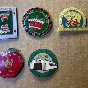 May include: Five metal enamel pins from the Pasadena Motorcycle Club. One rectangular playing card pin, two circular poker run pins, one arch-shaped poker run pin, and one apple-shaped pin. Each features text, dates, and graphic illustrations.