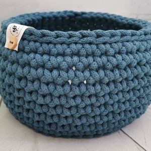 May include: A round, blue crochet basket with a textured stitch pattern. A small white tag with a wooden button is attached to the side. The basket is made of a thick, soft yarn and is suitable for storage.