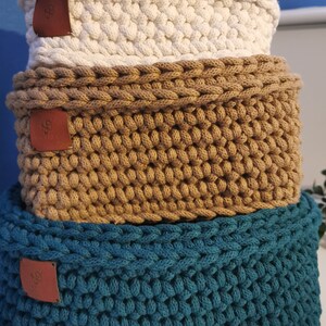 May include: Three stacked, hand-crocheted storage baskets in cream, tan, and teal. Each basket has a rectangular shape and a textured, woven appearance. Small, brown leather tags are attached to the front of each basket.