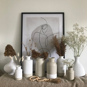 May include: A collection of seven ceramic vases in various shapes and sizes, with a white and beige color scheme. The vases are arranged on a brown textured surface, with dried flowers and a wooden bead garland in front of them. A framed abstract print with a black frame hangs on the wall behind the vases.