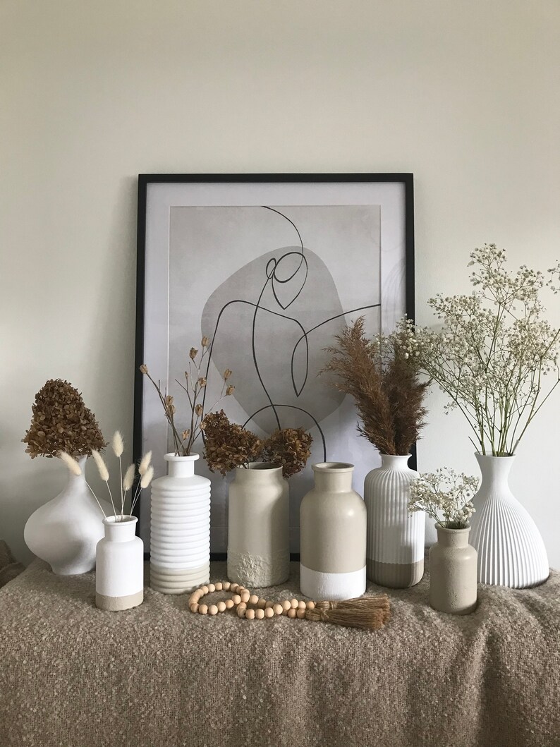 May include: A collection of seven ceramic vases in various shapes and sizes, with a white and beige color scheme. The vases are arranged on a brown textured surface, with dried flowers and a wooden bead garland in front of them. A framed abstract print with a black frame hangs on the wall behind the vases.