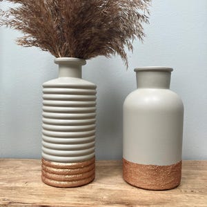 May include: Two decorative vases in a neutral colour scheme. One vase has horizontal ridges and the other is smooth. Both vases have a copper-coloured textured base. The vase on the left holds dried brown ornamental grass.