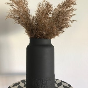 May include: A black vase with a textured bottom, filled with dried pampas grass.