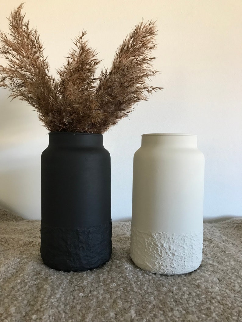 May include: Two ceramic vases, one black and one white, with a textured bottom. Both vases are filled with dried pampas grass.