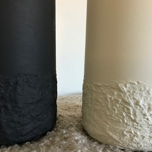 May include: Two ceramic vases, one black and one white, with a textured, rough finish. The vases are sitting on a light brown surface.