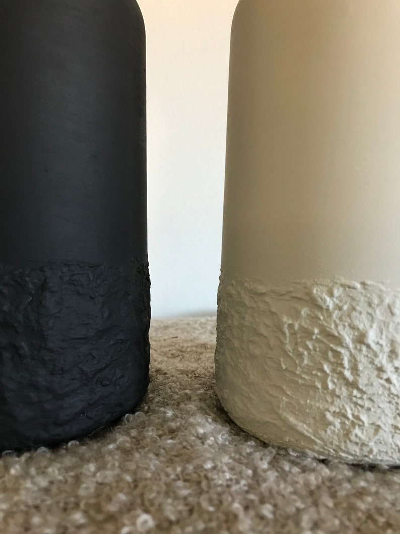May include: Two ceramic vases, one black and one white, with a textured, rough finish. The vases are sitting on a light brown surface.