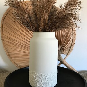 May include: White ceramic vase with a textured bottom, filled with dried pampas grass. The vase is sitting on a black tray.