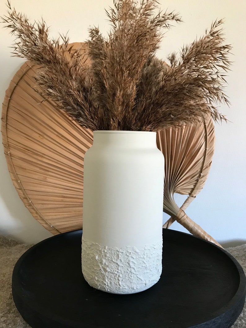 May include: White ceramic vase with a textured bottom, filled with dried pampas grass. The vase is sitting on a black tray.