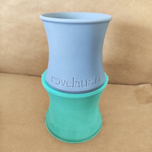May include: A two-piece ceramic vase in a light blue and teal color scheme. The top section is light blue with the word "revelation" etched into the surface. The bottom section is teal. The vase has a unique hourglass shape.