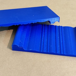 Può includere: Due oggetti blu brillante stampati in 3D. Uno è un blocco rettangolare con un bordo curvo e l'altro è un pezzo abbinato con una superficie scanalata e un bordo curvo. Gli oggetti poggiano su una superficie beige.