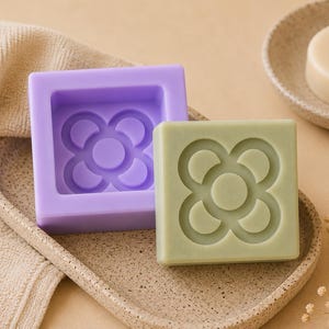 Pode incluir: Um sabonete quadrado verde claro com um desenho floral está ao lado de um molde de sabão roxo com o mesmo desenho. Uma barra de sabão redonda está em um prato pequeno. Os itens estão em uma bandeja salpicada.