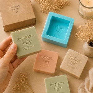 Pode incluir: Uma variedade de sabonetes quadrados em várias cores, cada um com a inscrição "custom text" e "handmade". Uma forma de sabonete azul e uma caixa castanha com um autocolante "handmade with love" também são visíveis. Uma vela acesa e flores secas completam a cena.
