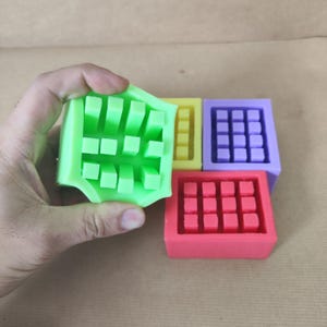 Peut inclure: Une main tenant un moule en silicone vert citron avec un motif en forme de grille de petites protubérances carrées. Trois autres moules en silicone jaunes, violets et rouges sont visibles en arrière-plan, chacun avec un agencement de grille différent. Les moules sont probablement destinés à l'artisanat ou à la pâtisserie.