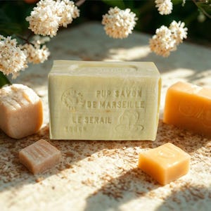 May include: A rectangular, pale yellow bar of soap with embossed text, "PUR SAVON DE MARSEILLE LE SERAIL 300GR", is the focal point. Several smaller, differently shaped soaps in shades of peach and orange surround it. White flowers are in the background.