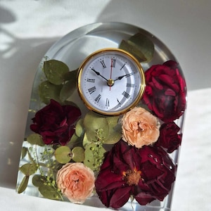 May include: A clear resin clock with dried flowers and greenery. The clock face has Roman numerals and gold hands.