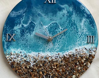 May include: A round clock with a blue and white resin ocean wave design. The clock face has Roman numerals and a silver hand. The bottom of the clock has a brown and white beach scene with small pebbles.