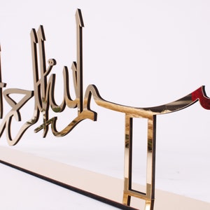 May include: A gold-colored metal wall decor with the Arabic phrase "Bismillah" in a decorative script. The decor is mounted on a stand with a rectangular base.
