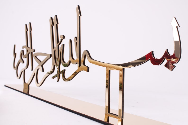 May include: A gold-colored metal wall decor with the Arabic phrase "Bismillah" in a decorative script. The decor is mounted on a stand with a rectangular base.