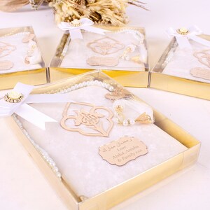 May include: Several gift boxes with gold-colored bases and clear lids. Each box contains a white prayer mat, a pearl-like rosary, and a decorative wooden accent. The boxes are tied with white ribbons and adorned with floral embellishments.