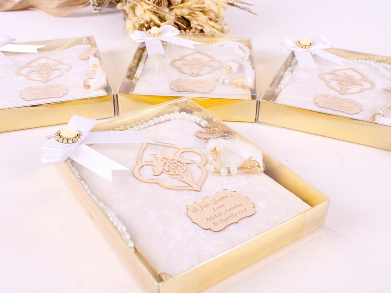 May include: Several gift boxes with gold-colored bases and clear lids. Each box contains a white prayer mat, a pearl-like rosary, and a decorative wooden accent. The boxes are tied with white ribbons and adorned with floral embellishments.